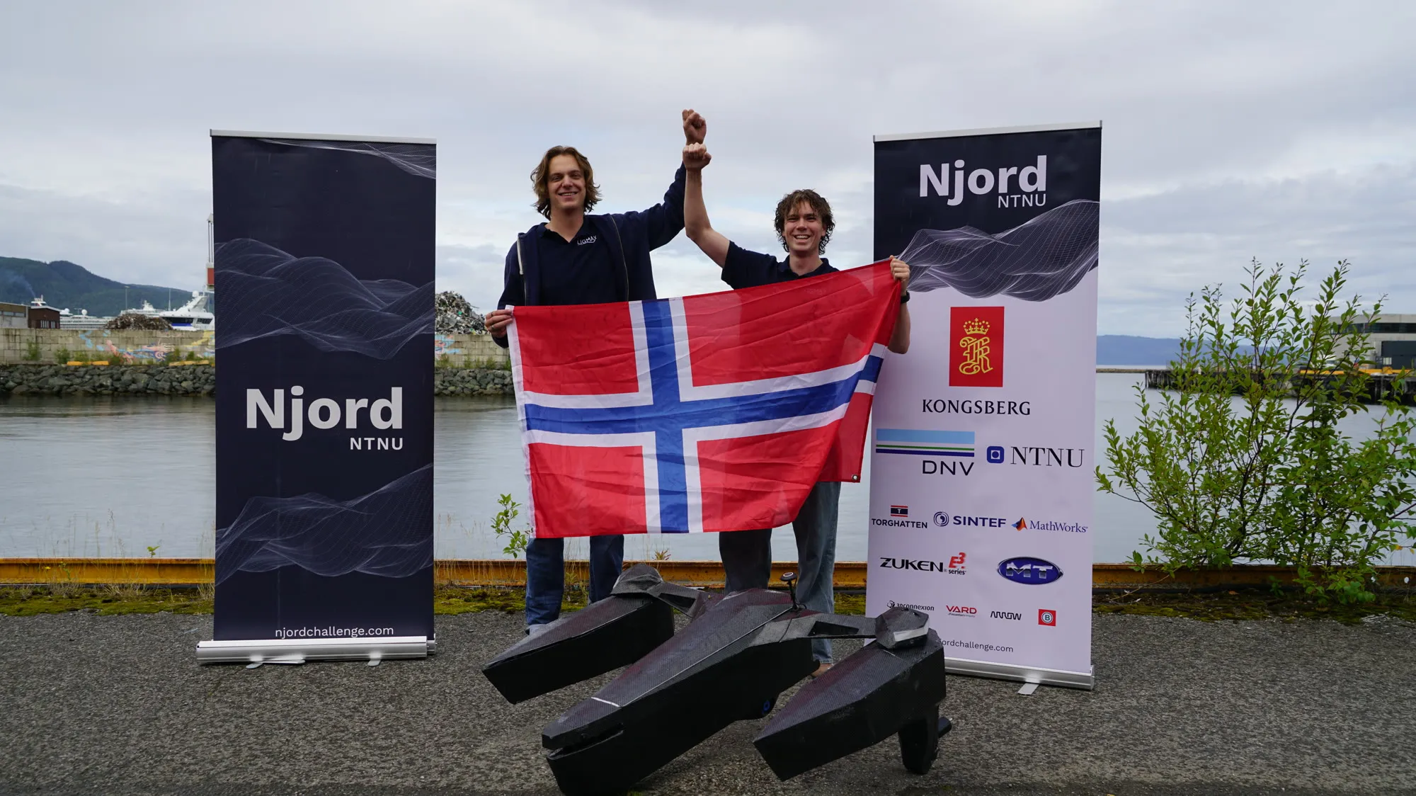Two Ligmax team members holding a Norwegian flag behind the vessel at the Njord 2026 competition site